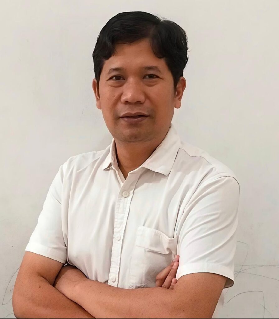 Man with short black hair wearing a white button down shirt arms crossed facing the camera against a pale wall
